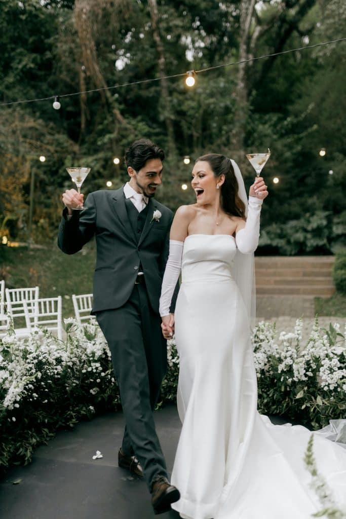 Bride and groom joyfully celebrating their wedding outdoors with drinks in a lush garden setting.